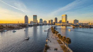 "Aerial view of Jacksonville, Florida featuring the St. Johns River, Main Street Bridge, and downtown skyline during a sunset in January, with people strolling along the Riverwalk and boats on the water."