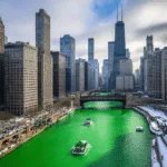 "Chicago skyline on St. Patrick's Day with green-dyed river, buildings in shadows, snow patches, people in green attire on riverwalk, and boats on the river"