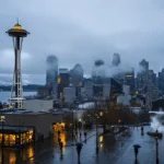 "Misty aerial view of Seattle's skyline with Space Needle emerging through clouds, city lights reflecting on Puget Sound, and Mount Rainier in distance"