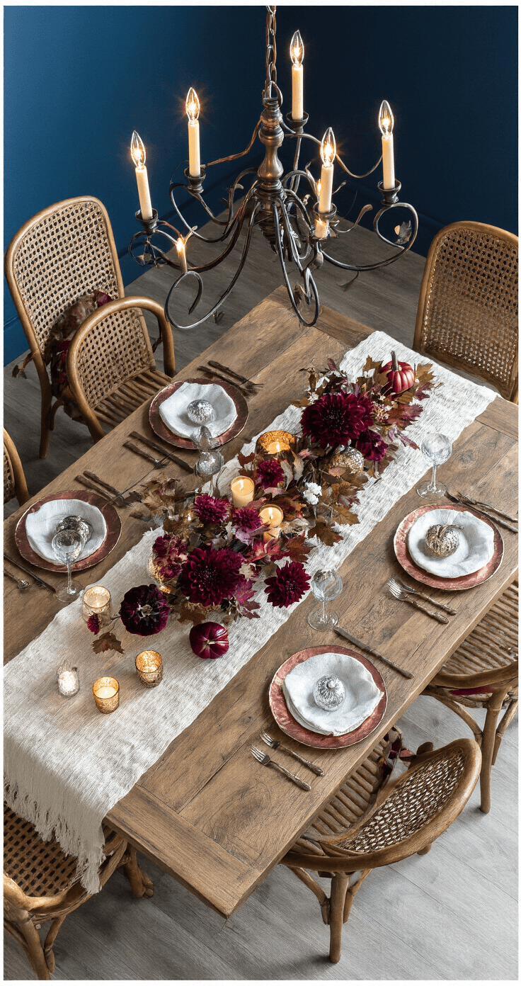 Cozy Fall Decor: Transform Your Home into an Autumn Haven Overhead view of a dramatic dining room tablescape at blue hour, featuring an 8ft reclaimed wood table with a textured ivory runner, mercury glass votives, jewel-toned velvet pumpkins, and an organic arrangement of burgundy dahlias, maple branches, and copper beech leaves. Vintage copper chargers reflect candlelight, and woven rattan chairs add texture, illuminated by a dramatic chandelier.