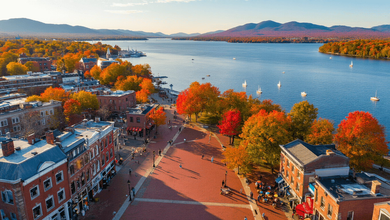 "Aerial view of Burlington, Vermont in autumn with Church Street Marketplace, ECHO Leahy Center, Lake Champlain, and Adirondack Mountains; street performers, outdoor cafés, and First Unitarian Universalist Society church present."