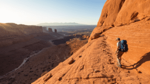 "Hiker traversing steep, slickrock trail at Arches National Park, with distant view of Delicate Arch, cairn markers and challenging elevation changes signifying trail difficulty."