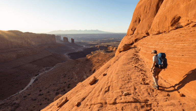 The Hidden Truth About Hiking to Delicate Arch That Nobody Tells You (And Why Your First Attempt Might Be Brutal) "Hiker traversing steep, slickrock trail at Arches National Park, with distant view of Delicate Arch, cairn markers and challenging elevation changes signifying trail difficulty."