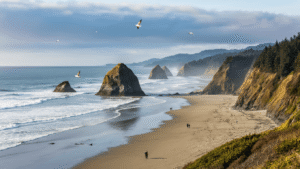 "Hikers walking along Oregon's coastline with Haystack Rock sea stacks, coastal rainforest, and morning lit waves in view, and Oregon Coast Trail markers in the distance."