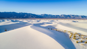 "Aerial view of White Sands National Park's white gypsum dunes under a blue sky, framed by mountains, with a road winding through and sparse vegetation providing contrast."